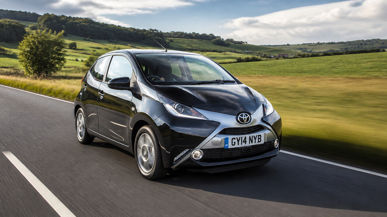 Carro Toyota preto a circular numa estrada asfaltada rodeada por campos verdes e árvores sob céu nublado.
