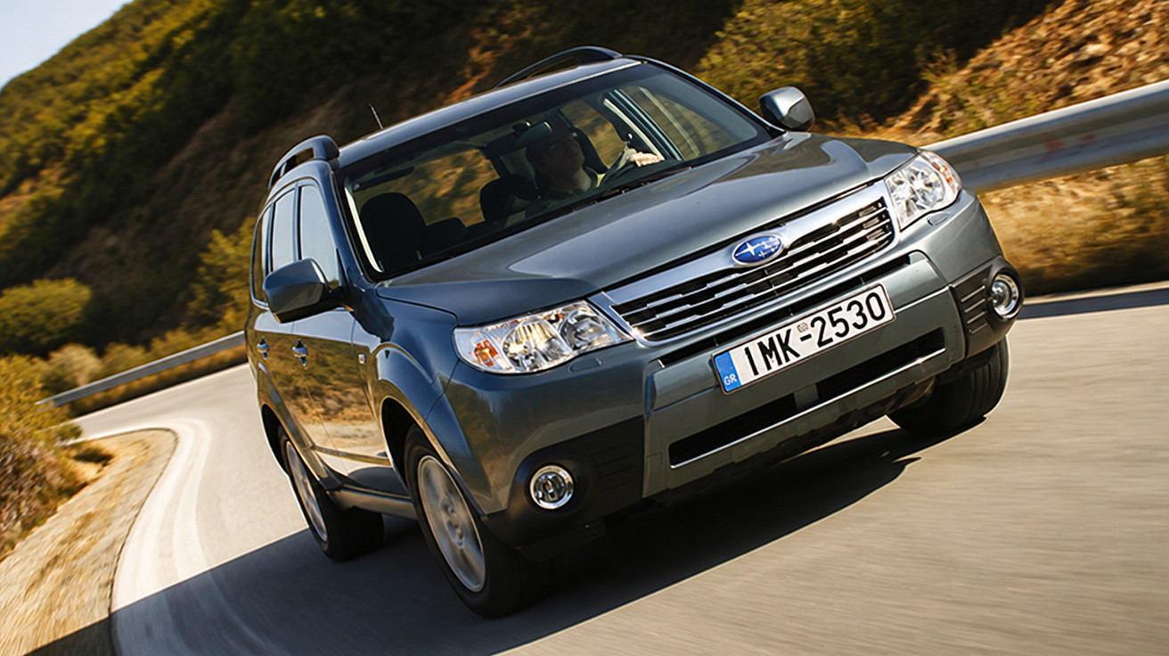 Carro Subaru cinzento a conduzir numa estrada sinuosa com proteções metálicas e vegetação ao lado.