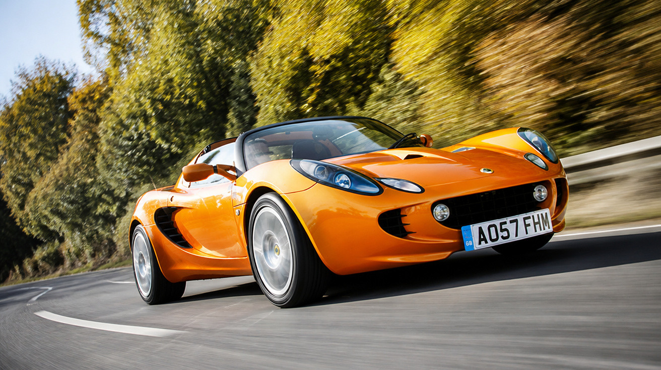 Carro desportivo descapotável laranja a circular numa estrada curva com árvores ao fundo.