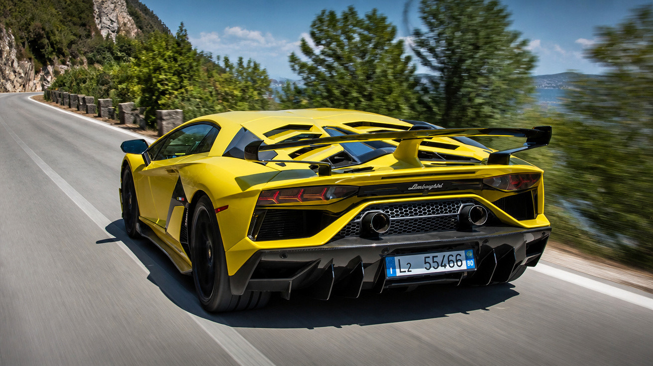 Carro desportivo amarelo Lamborghini a acelerar numa estrada sinuosa junto a rochas e árvores.
