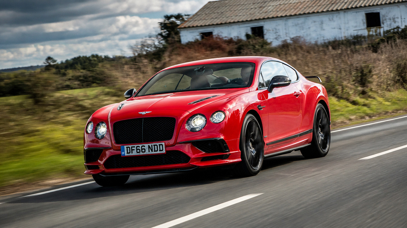 Carro desportivo vermelho Bentley a circular numa estrada rural com casa ao fundo durante dia nublado.